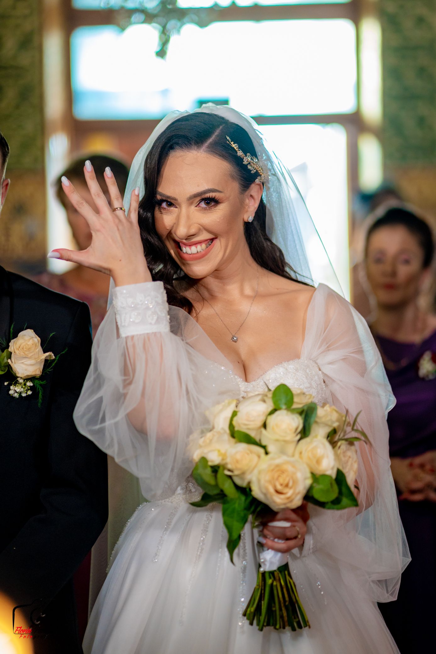 Joyful bride in white lace dress waves while holding cream rose bouquet during ceremony.
