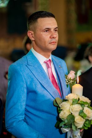 Groomsman in light blue suit with pink tie sits beside floral arrangements and candles.