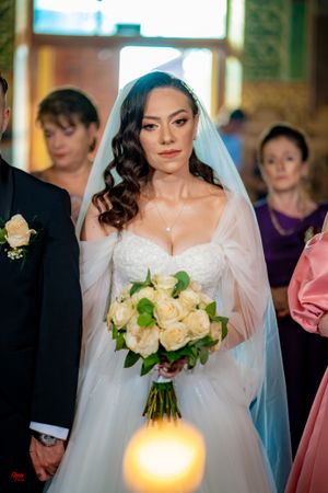 Bride in white lace gown holds cream rose bouquet during wedding ceremony.