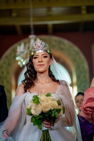 Bride wearing an ornate crown and veil holds a bouquet of cream roses during the ceremony.