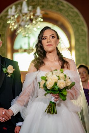 A bride in a white gown holds a cream rose bouquet under an ornate archway.