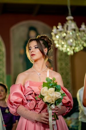 Elegant bridesmaid in a pink off-shoulder gown with dramatic puff sleeves holds a bouquet under a crystal chandelier.