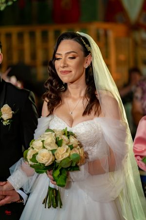 Bride in white lace gown and veil smiles while holding cream rose bouquet during ceremony.