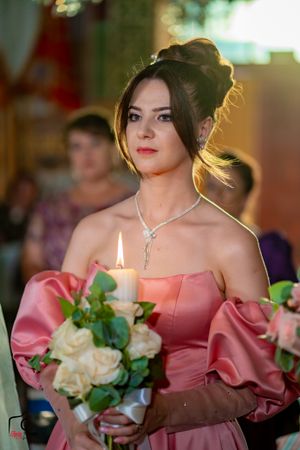 Bridesmaid in coral off-shoulder dress holds flowers and candle during evening ceremony.