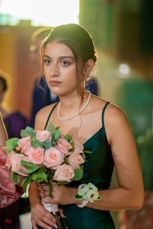 Bridesmaid in emerald green dress holds pink rose bouquet with pearl accessories.