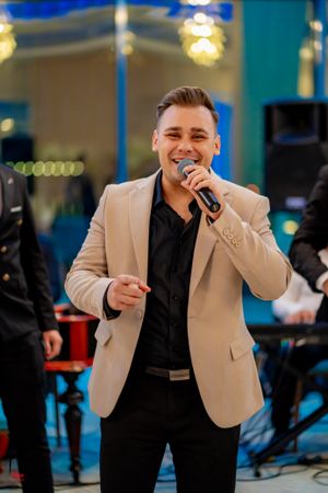 Performer in cream blazer and black pants singing with a microphone at an indoor event with blue lighting.