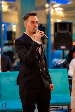 Entertainment performer singing into microphone wearing dark suit and glasses in blue-lit venue.