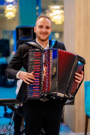 Musician playing red and black accordion while wearing formal attire at indoor event.