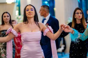 Women in pink and red dresses dance in formation at an elegant indoor event.