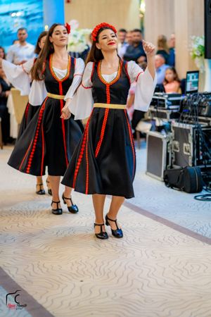 Folk dancers in traditional black and red costumes performing at an indoor venue.