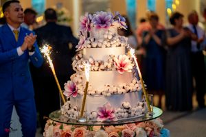 Elegant white wedding cake decorated with purple flowers and sparklers glowing in dim reception lighting.