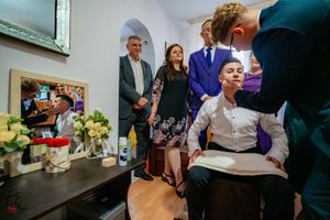 A group watches an interaction in a home setting with flowers and mirrors visible in the background.