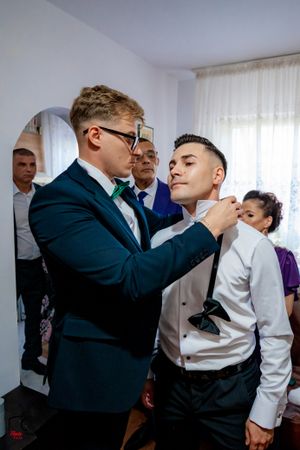 A groomsman helps adjust another's formal attire in a bright room while others look on during wedding preparations.