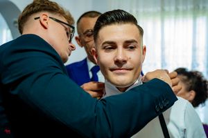Wedding preparation scene showing careful adjustment of formal attire in natural lighting.
