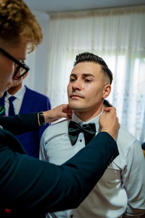A person in formal attire having their black bowtie adjusted during wedding preparations.