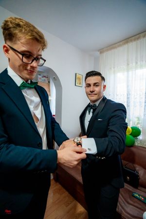 Two people in formal suits helping each other with cufflinks in a bright room.