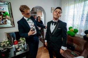 Two groomsmen in formal black tuxedos share a lighthearted moment while getting ready in a living room.