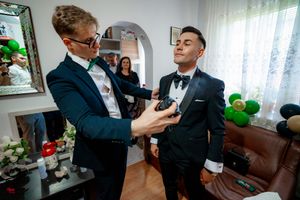 Groomsmen in black tuxedos help each other with formal attire preparations in a home setting.