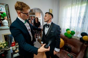 Two men in formal attire share laughter while adjusting bowties before an event.