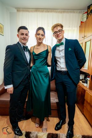 Three people in formal attire pose together, with a woman in an elegant green dress between two men in tuxedos.