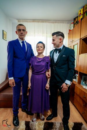 Family portrait with three people in formal wear standing against wooden furniture and white curtains.
