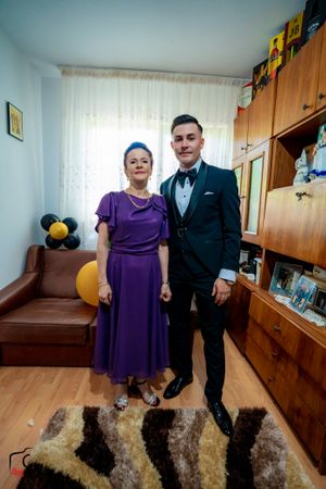 Woman in purple dress poses with man in black tuxedo against wooden furniture and decorative balloons.