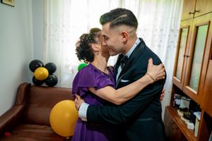 Dancers in formal wear embrace during a tender moment by a window.