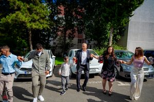 Wedding guests gather outside on a sunny day near parked cars and trees.