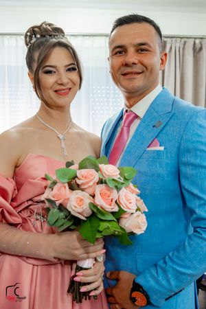 A couple poses with pink roses against cream curtains in formal attire.