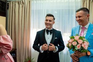 Two men in formal wear, one in black tuxedo and one in light blue suit holding pink roses.