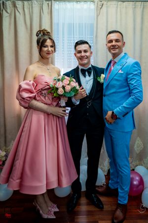 Wedding party in pink dresses and suits poses together against cream curtain backdrop.