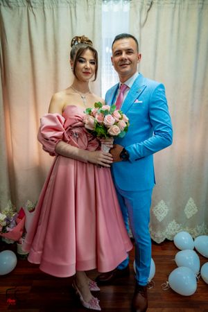 A couple poses in formal attire with a pink dress and bright blue suit holding a bouquet of pink roses.