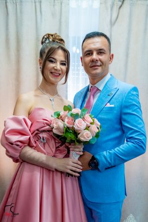 A couple in pink and blue formal wear poses with a bouquet against a light backdrop.