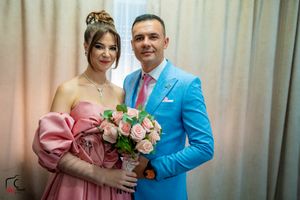 A couple smiles while posing with pink roses against a curtained backdrop.