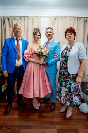 Family portrait with couples in blue suits and pink dress against beige curtains.