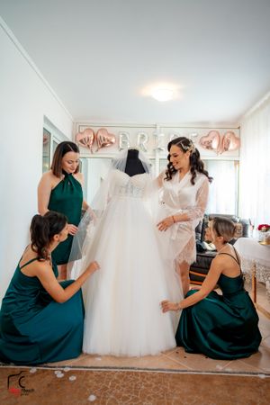 Bridesmaids in emerald green dresses admire a white wedding gown displayed in a bridal preparation room.