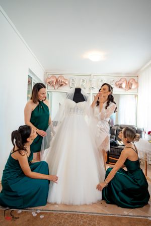 Women in dark green gowns gather around a white tulle wedding dress in a bright room.