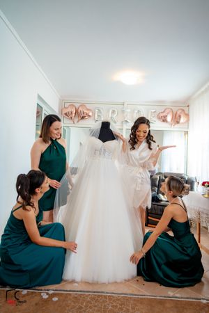 Women in matching emerald dresses gather around a white bridal gown in preparation room.