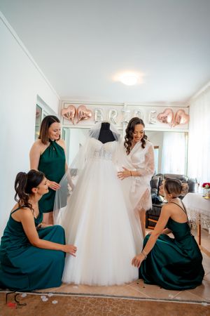 Bridesmaids in dark green attire share candid moments near a displayed wedding dress.