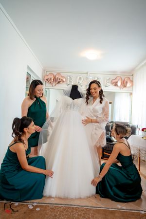 Women in coordinated green gowns admire and interact around a white wedding dress.