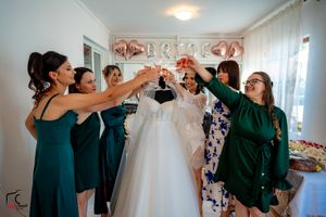Wedding party members in emerald dresses gather around a hanging white wedding gown in a bright room.