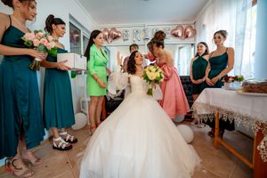 Bridesmaids in teal and coral dresses help prepare for the wedding celebration in a sunlit room.