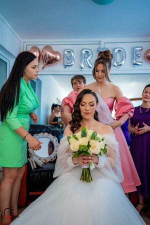Bride holding yellow flower bouquet while surrounded by bridesmaids in a getting-ready room.
