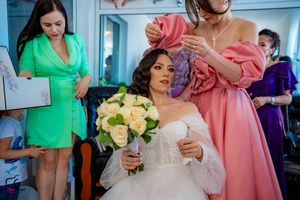 Wedding preparations with bridesmaids in colorful dresses helping with final touches while holding a cream rose bouquet.
