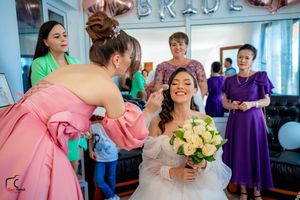 A bride in a white dress holds yellow roses while getting her makeup done, surrounded by attendants in pink and purple.