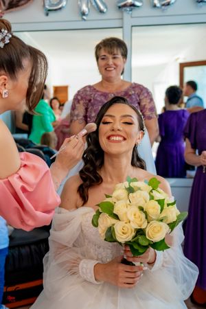 A joyful bride holding cream roses gets her makeup applied while wearing an off-shoulder wedding gown.