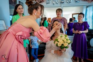 A makeup artist in a pink dress touches up a laughing bride's face during wedding preparations.
