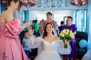 A bride in a white ballgown holds yellow roses while getting final makeup touches under balloon decorations.