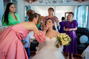 Bridal preparations show a makeup artist in coral pink working on a bride's face amid wedding party members.