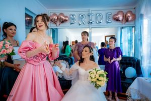 Wedding party scene with bridesmaids in purple dresses and pink decor in a blue-walled room.